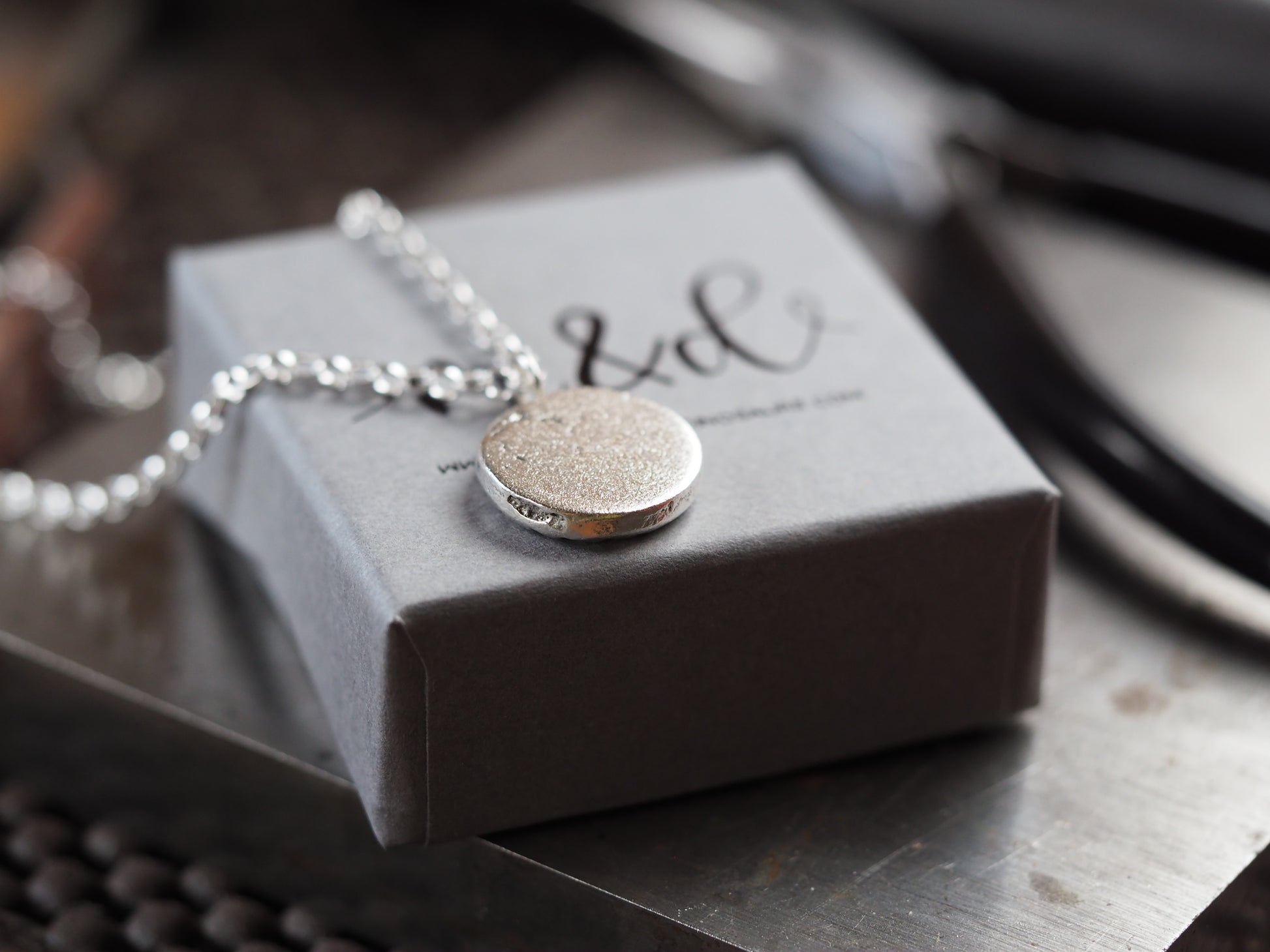 Silver necklace with a round pendant on a grey box with 'M&D' branding.