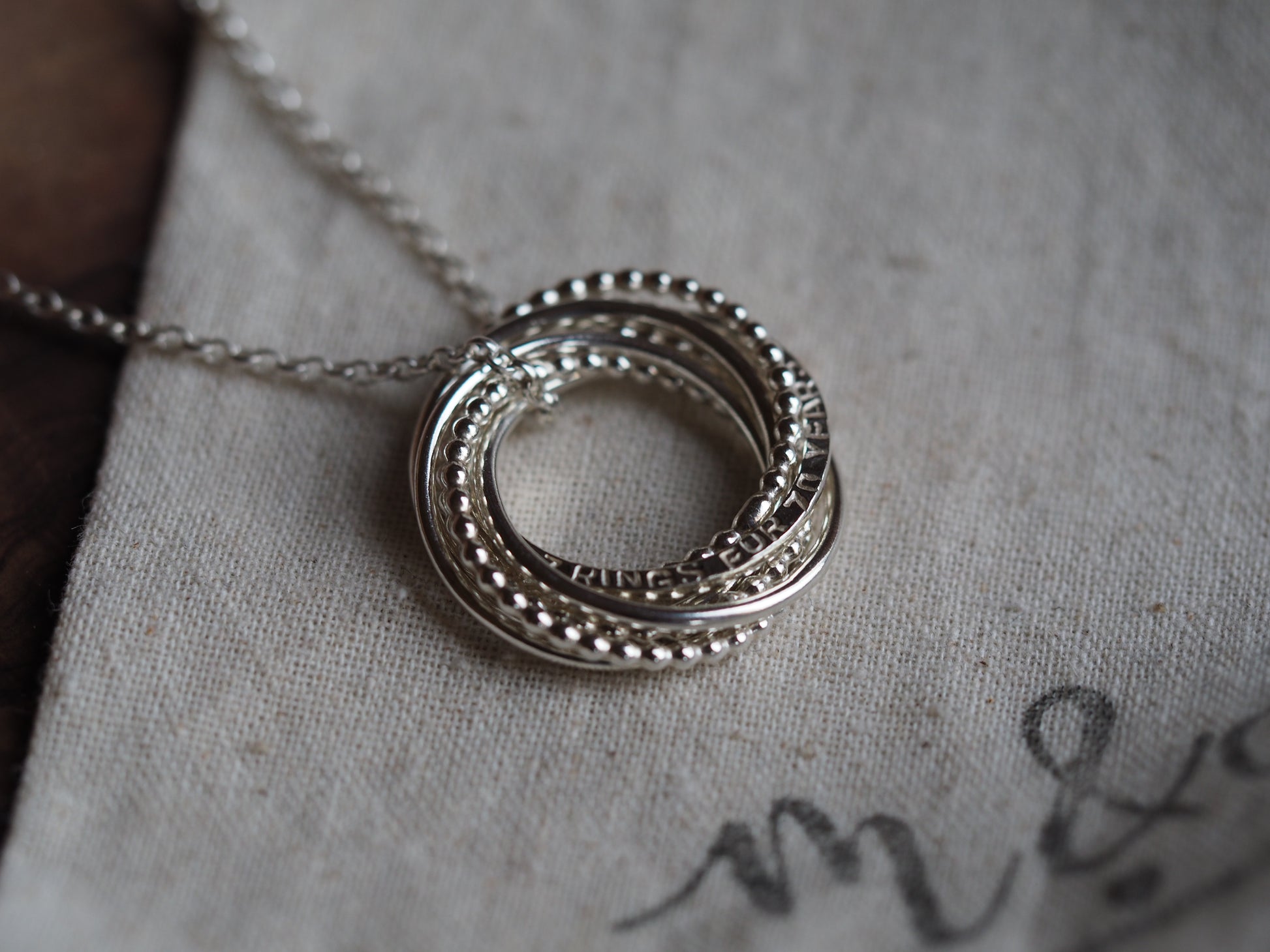A silver interlocking ring necklace laid out on a fabric surface, with the ring part in focus showing a textured design.