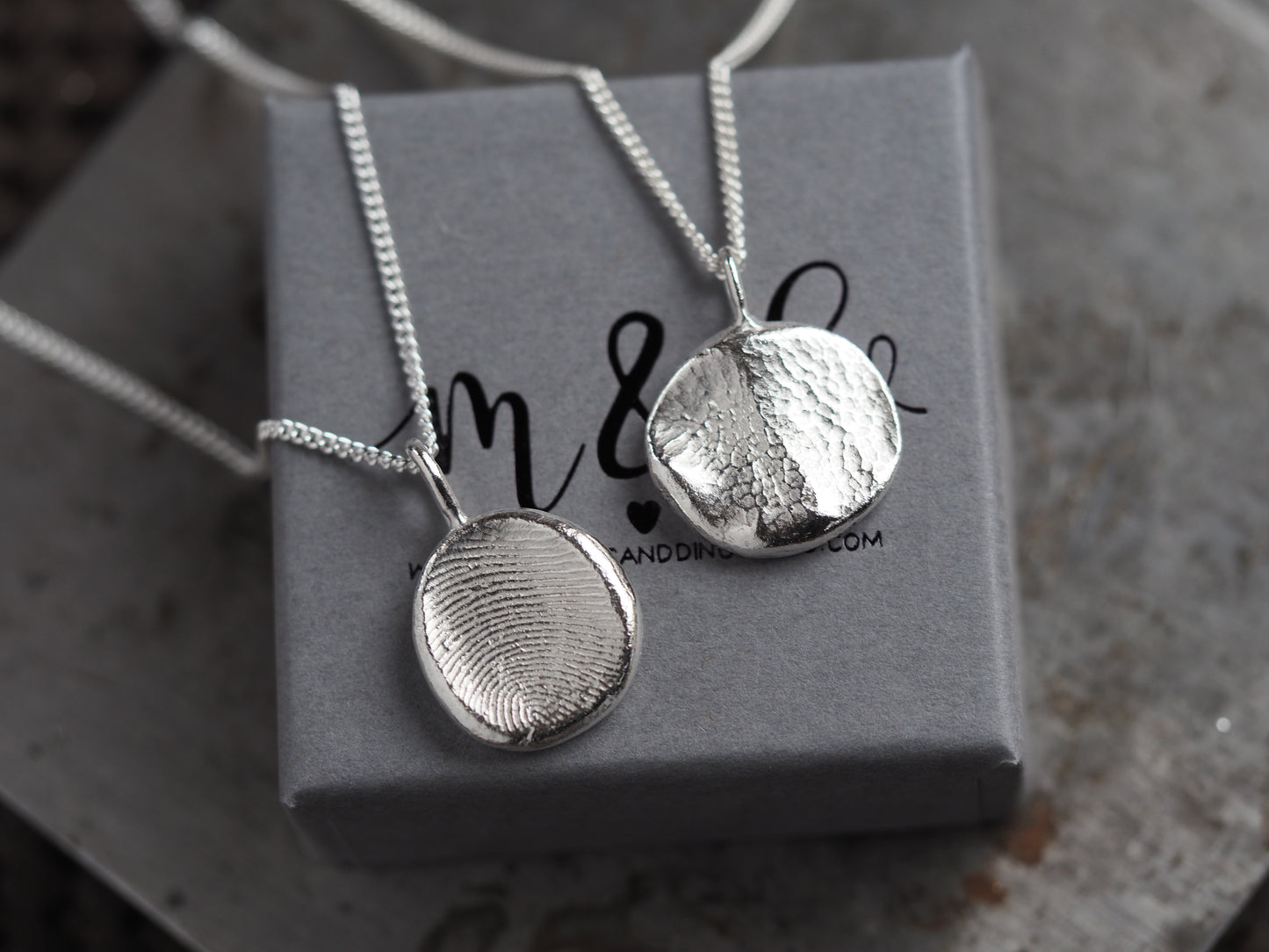 Two silver pendants on chains resting on a gray box with 'm&d' branding.