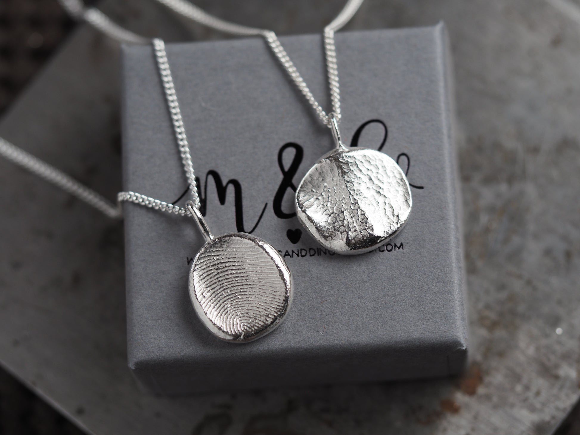 Two silver pendants on chains resting on a gray box with 'm&d' branding.